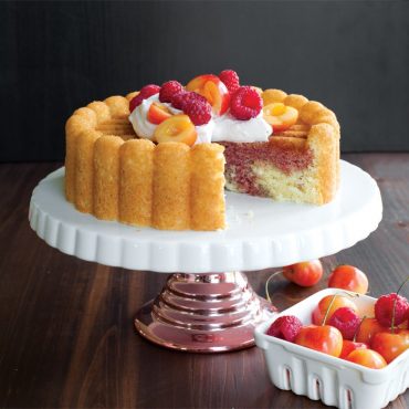 Marbled cherry charlotte cake with slice removed, topped with whipped cream and raspberries on white cake stand