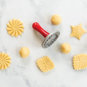 Three Nordic Ware Pretty Pleated cookie stamps with floral designs pressed into raw cookie dough balls on floured surface