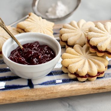 Round sandwich cookies with decorative stamped patterns, filled with red berry jam and dusted with powdered sugar