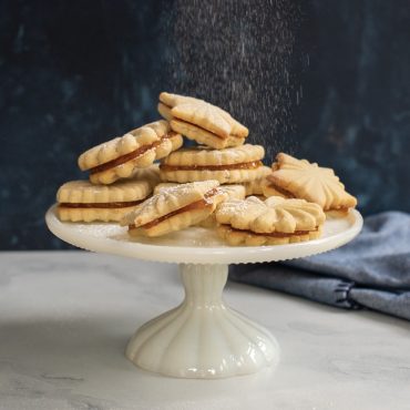 Sandwich cookies with decorative stamped patterns filled with apricot jam, dusted with powdered sugar on serving plate