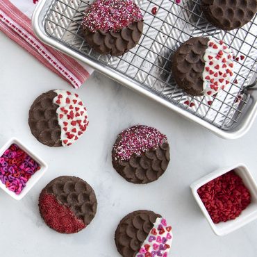 Round cookies with decorative stamped patterns, half dipped in white chocolate and colorful sprinkles on baking sheet