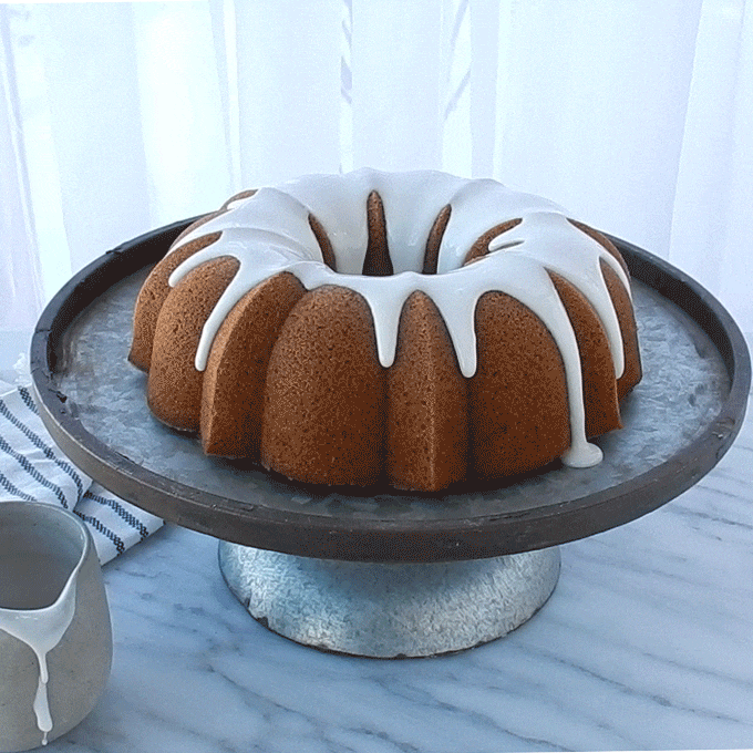 Hand drizzling white glaze over golden bundt cake on glass pedestal stand, cake showing detailed fluted ridges