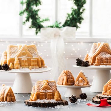 Baked vanilla house-shaped bundt cakes on white stands, dusted with powdered sugar, decorated with pine cones