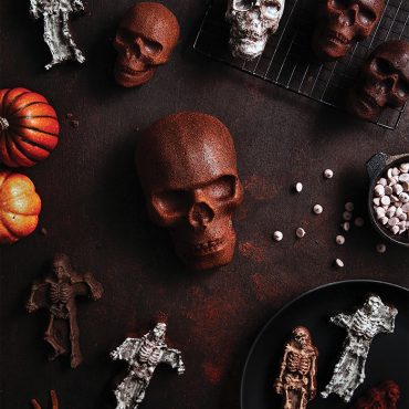 Nordic Ware Halloween skull and skeleton cakelets with orange frosting displayed alongside decorated cakes