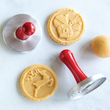 Red handled Nordic Ware cookie stamps with stamped sugar cookies showing reindeer and holly designs