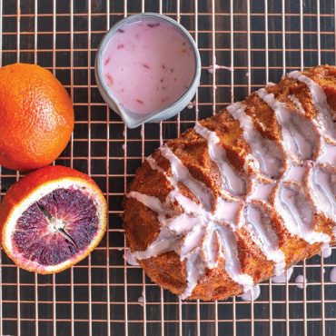 Sliced blood orange loaf cake with pink glaze drizzle on cooling rack, bowl of glaze and fresh oranges alongside