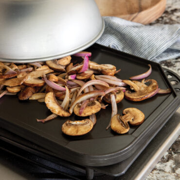 Sautéed mushrooms and red onions on square griddle pan with Nordic Ware aluminum high-dome lid partially covering
