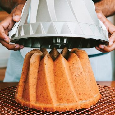 Hands lifting Nordic Ware Kugelhopf bundt pan revealing golden baked cake with distinctive fluted ridges