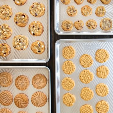 Four Nordic Ware aluminum Prism baking sheets with textured surface, displaying freshly baked cookies including chocolate chip and sugar cookies