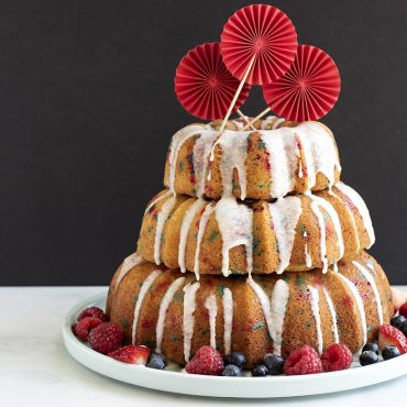 Three-tiered golden bundt cakes with white glaze drizzle, garnished with berries and red paper fans