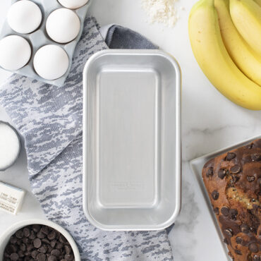Nordic Ware natural aluminum 1-pound loaf pan with baking ingredients including flour, eggs, and butter arranged around it