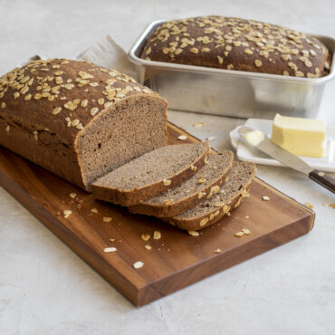 Sliced whole wheat oatmeal bread on cutting board with Nordic Ware aluminum 1-pound loaf pan in background
