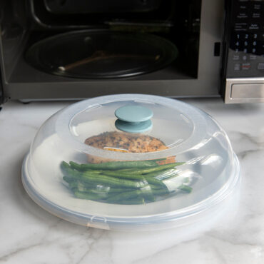 Clear Nordic Ware Up & Away spatter cover over white plate with cooked chicken and green beans, microwave in background