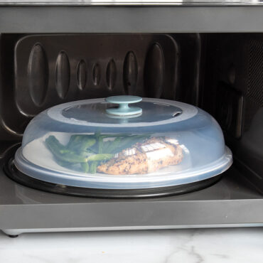 Nordic Ware clear plastic spatter cover over white plate with cooked chicken and green beans in microwave