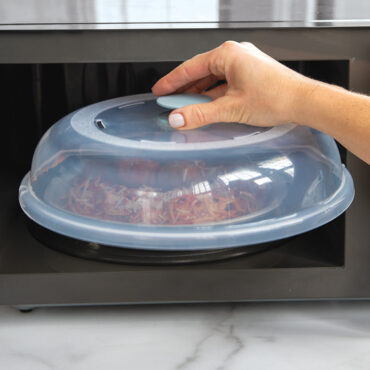 Hand lifting Nordic Ware Up & Away clear plastic spatter cover from microwave containing plate of spaghetti