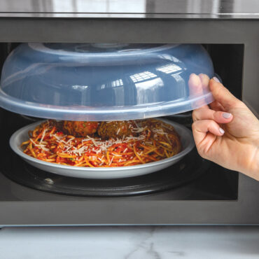 Hand pulling Nordic Ware Up & Away spatter cover from microwave above plate of cooked spaghetti