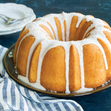Golden glazed bundt cake with decorative fluted ridges on white cake plate, baked in Nordic Ware pan