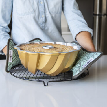 Woman using Nordic Ware bundt-shaped hot pads to transfer golden bundt cake in aluminum pan to cooling rack
