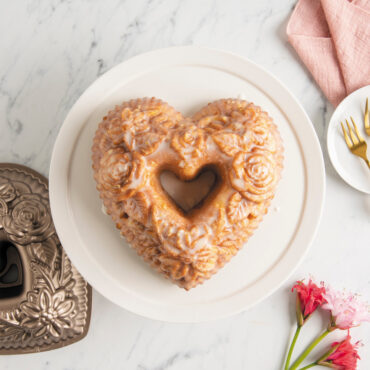 Overhead of glazed Floral Heart Bundt on white cake stand with a plate of diced strawberries, flowers, and pan