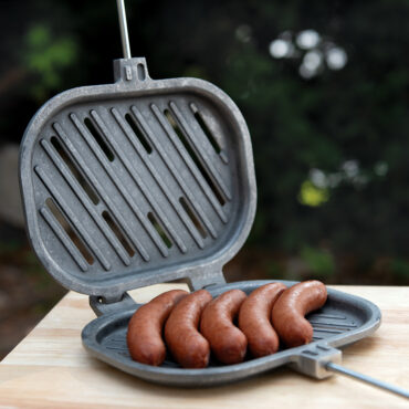 Cast aluminum campfire griller opened flat showing hinged cooking basket with grilled sausages inside