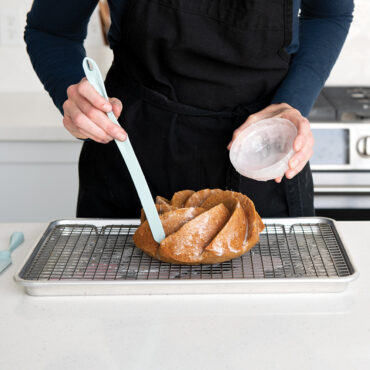 Nordic Ware flexible blade tool applying white glaze to golden bundt cake surface