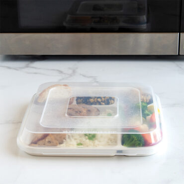 Nordic Ware aluminum divided dinner tray with clear lid, containing leftover food portions, positioned in front of microwave