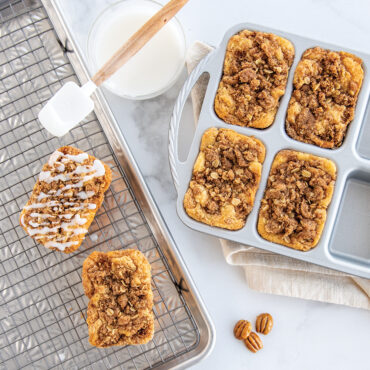 Six golden mini loaves with nut topping in Nordic Ware aluminum ProCast mini loaf pan, glazing drizzle nearby