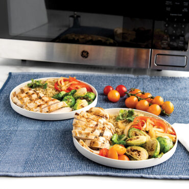 Nordic Ware white 8-inch lunch plate with food serving, displayed on blue placemat with microwave in background