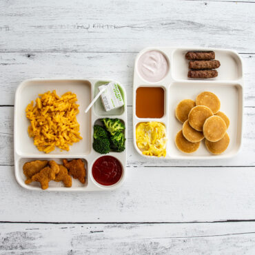 Two white Nordic Ware divided meal trays with breakfast and lunch foods in separate compartments