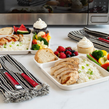 Two Nordic Ware divided meal trays with compartments filled with chicken, rice, vegetables, fruit, and cupcakes near microwave