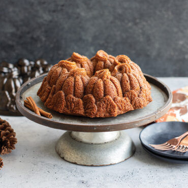 Golden bundt cake with pumpkin-shaped sections on white cake stand, Nordic Ware Pumpkin Patch pan visible behind