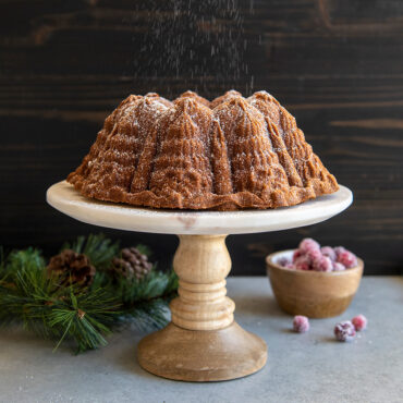 Golden bundt cake with decorative ridged pattern on white cake stand, garnished with pine sprigs and red cranberries