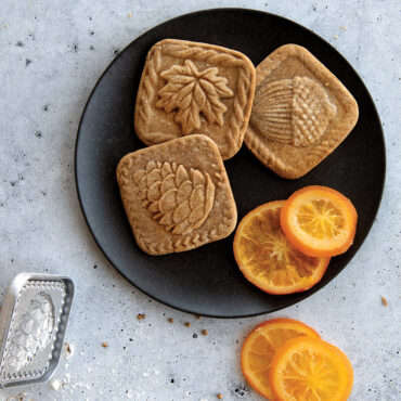 Decorated springerle cookies with forest motifs arranged on black plate beside orange slices, showing stamped designs