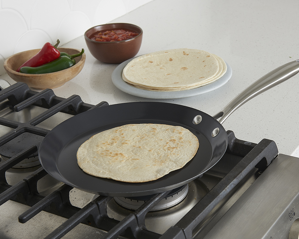 Nordic Ware's French Steel Crepe Pan on a gas burning stove top heating a flour tortilla until slightly browned. Next to the stove, there is a plate of cold tortillas, a bowl of salsa, and a bowl of hot peppers.