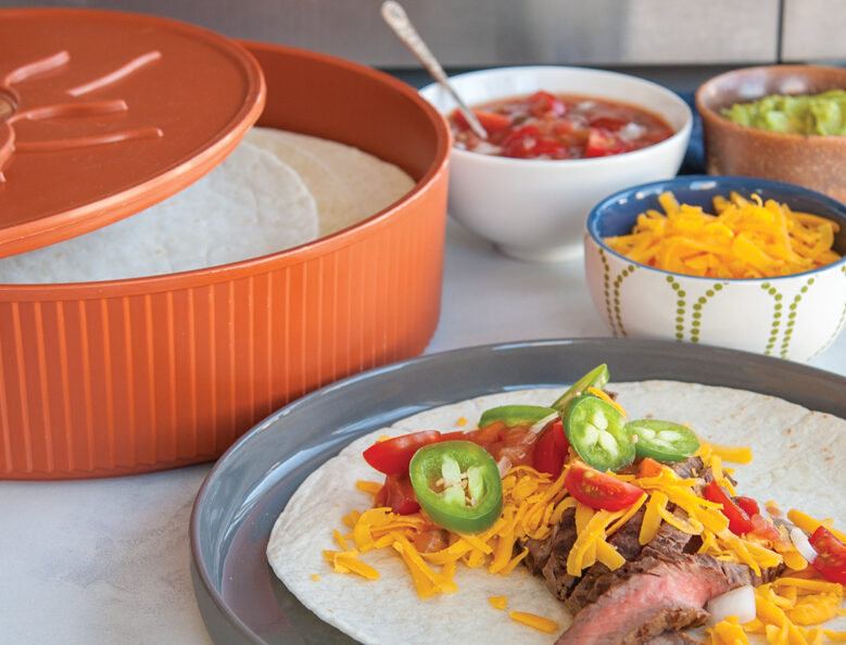 Nordic Ware's Tortilla and Pancake Warmer filled with warm tortillas next to a gray plate with a steak taco topped with cheese, tomatoes, and peppers. The to the plate, there is a bowl of cheddar cheese, a bowl of salsa, and a bowl of guacamole.