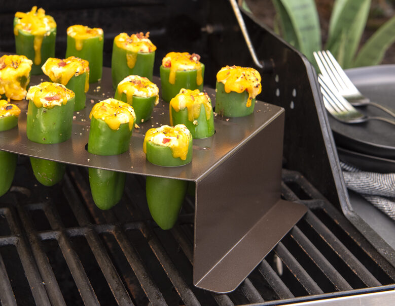 Nordic Ware's Jalapeno Roaster filled with stuffed jalapenos topped with cheddar cheese. Next to the grill, there are plates and folks to serve the jalapenos on an a grey kitchen towel to clean up any messes.
