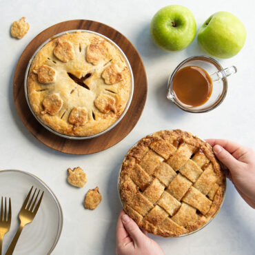 Two golden apple pies baked in Nordic Ware aluminum mini pie pans with hands lifting one pie, apples nearby