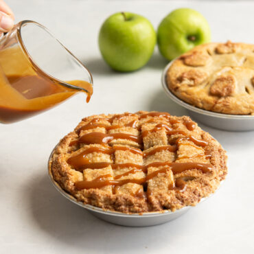 Hand drizzling caramel sauce over golden apple pie in Nordic Ware aluminum mini pie pan, second pie visible behind