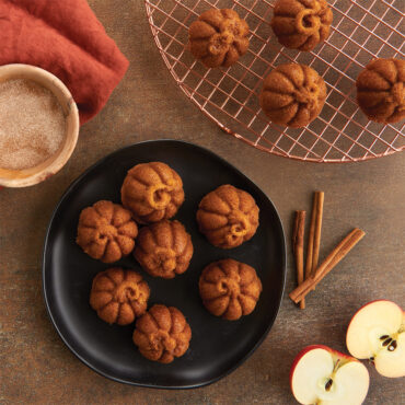 Six golden pumpkin-shaped cakelets on white plate with copper cooling rack, cinnamon sticks and apples nearby