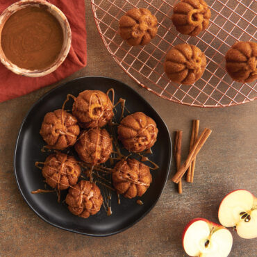 Six golden pumpkin-shaped cakelets with caramel drizzle on white plate, copper cooling rack, apples and cinnamon sticks