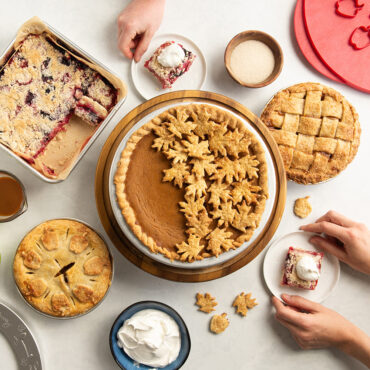 Baked pies and bars from Nordic Ware mini pie pans: whole pumpkin pie, two mini apple pies, and sliced pie bars with hands reaching for pieces