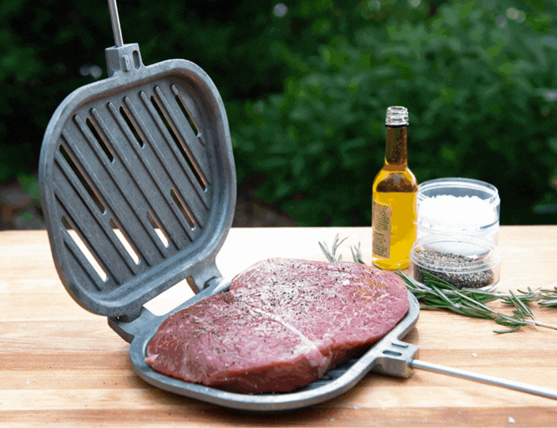 Campfire Griller Steps Gif This gif features the step-by-step process of using the Campfire Griller. The first image features an uncooked steak being seasoned on inside of the preheated griller on top of a wooden table. The second image features the closed Campfire Griller cooking the steak over an open campfire. The final image features the fully cooked steak inside of the Campfire Griller next to the campfire. In this image, the Campfire Griller is open to reveal the grill lines made by the Griller.