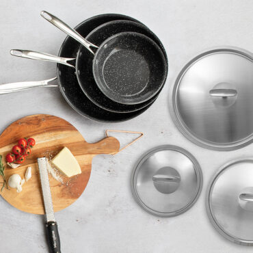 Nordic Ware Basalt 6-piece nonstick skillet set with glass lids, dark gray coating, displayed with cheese and sliced tomatoes