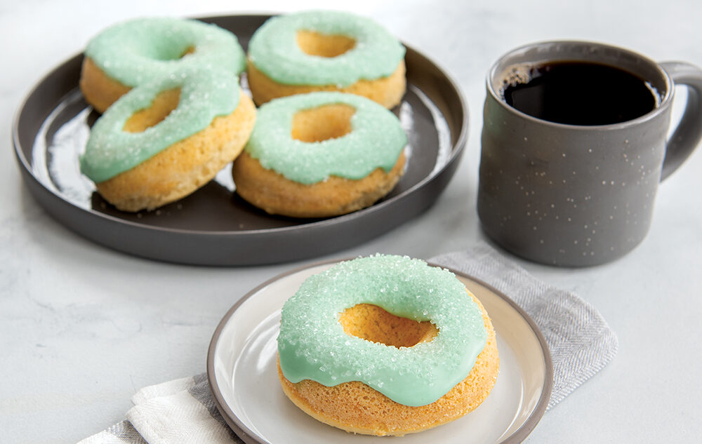 Five baked vanilla donuts glazed in a teal frosting. One donut in on a cream colored plate in the front and the other four are on a grey plate in the back of the image. There is also a cup of coffee next to both plates in a grey mug.