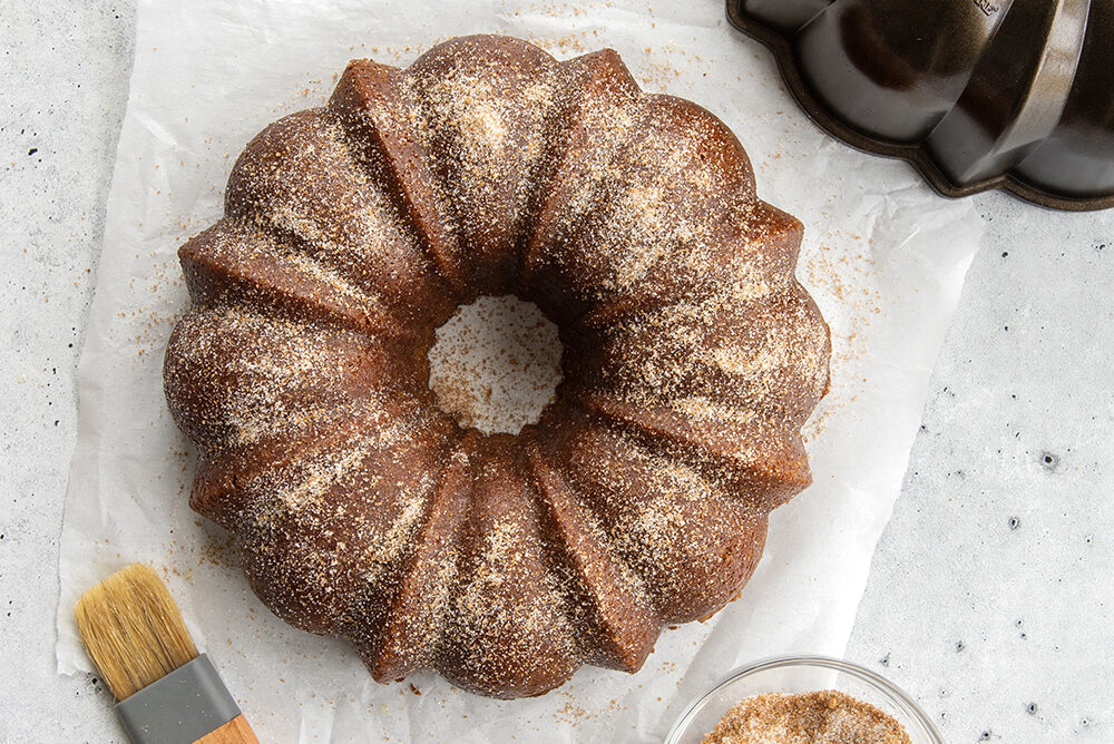 Cinnamon Spice Donut Bundt Cake made in a Bronze Original Cast Bundt Pan. The cake is covered in cinnamon sugar and is placed on a piece of parchment paper. Around the cake, there is the Bundt pan it was baked in, a bowl of cinnamon sugar, and a pastry brush used to brush the cake with butter.
