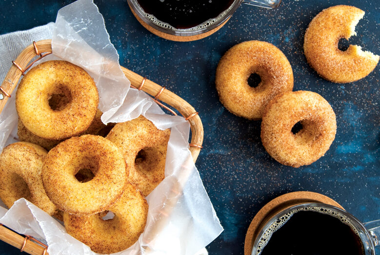 Cinnamon sugar coated mini donuts. Three of the donuts are on the table and the rest are in a wire basket lined with parchment paper. There are also two cups of coffee on the table next to the donuts.