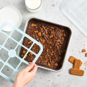 Hand holding Nordic Ware brownie cutter over aluminum pan of golden baked brownies, with lid nearby