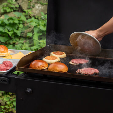 Nordic Ware cast iron grill press flattening burger patties on outdoor grill grates with hamburger buns nearby