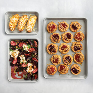 Nordic Ware 3-piece nonstick sheet pan set with baked cookies and roasted vegetables on gray background