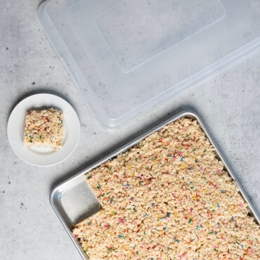 Nordic Ware aluminum sheet pan with lid containing golden rice crispy treats, one square piece plated beside